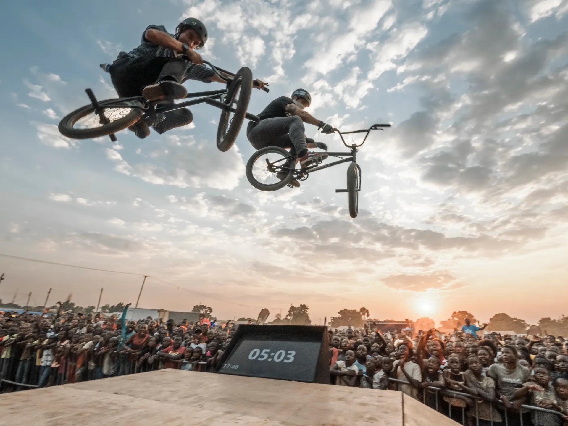 Two StuntDudes riders airborne at once over a huge crowd at sunset